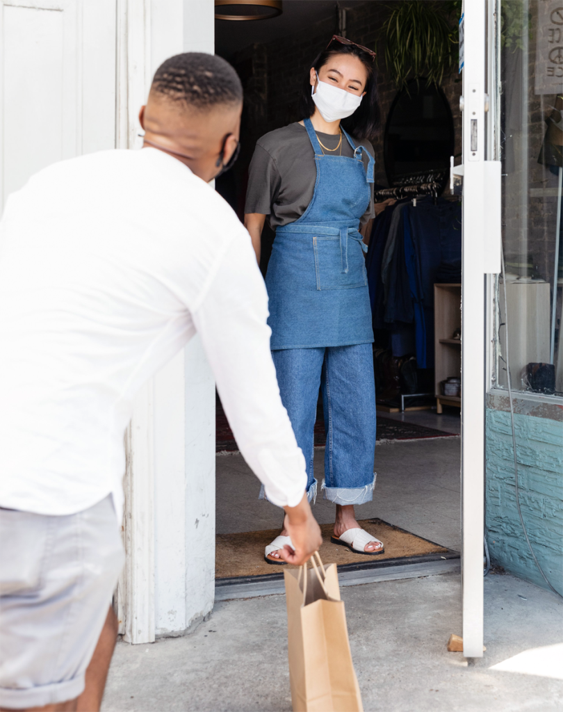 A masked but obviously smiling store employee (woman, Asian decent) handing off a curbside pickup order to a customer. The customer is a black man with his back to the camera.